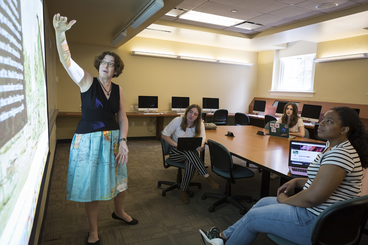A professor points at a project screen as three seated students watch